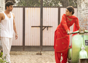 Pavia Sidhu as Jassi standing next to a green moped looking at Yugam Sood as Mithu standing outside in the movie Dear Jassi.