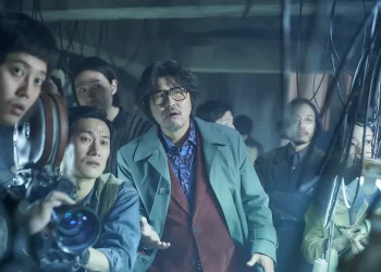 Song Kang-ho as Kim Ki-yeol standing in front of a group with his hand out stretched in the movie Cobweb.