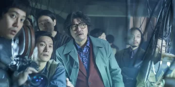 Song Kang-ho as Kim Ki-yeol standing in front of a group with his hand out stretched in the movie Cobweb.