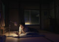 Koji Yakusho as Hirayama laying on a tatami reading a book by lamp in the movie Perfect Days.