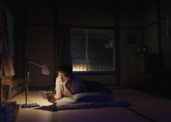 Koji Yakusho as Hirayama laying on a tatami reading a book by lamp in the movie Perfect Days.