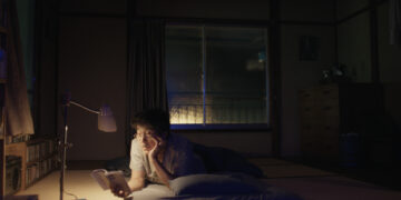 Koji Yakusho as Hirayama laying on a tatami reading a book by lamp in the movie Perfect Days.