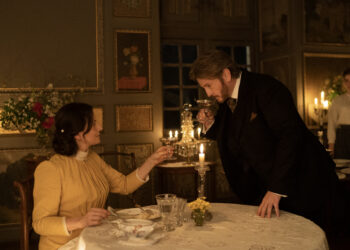 Juliette Binoche as Eugénie seated at the dinner table and Benoît Magimel as Dodin Bouffant standing next to her holding her hand in the movie The Taste of Things.