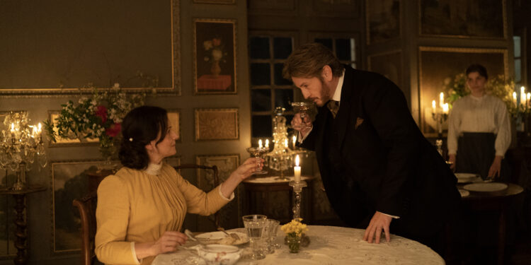 Juliette Binoche as Eugénie seated at the dinner table and Benoît Magimel as Dodin Bouffant standing next to her holding her hand in the movie The Taste of Things.