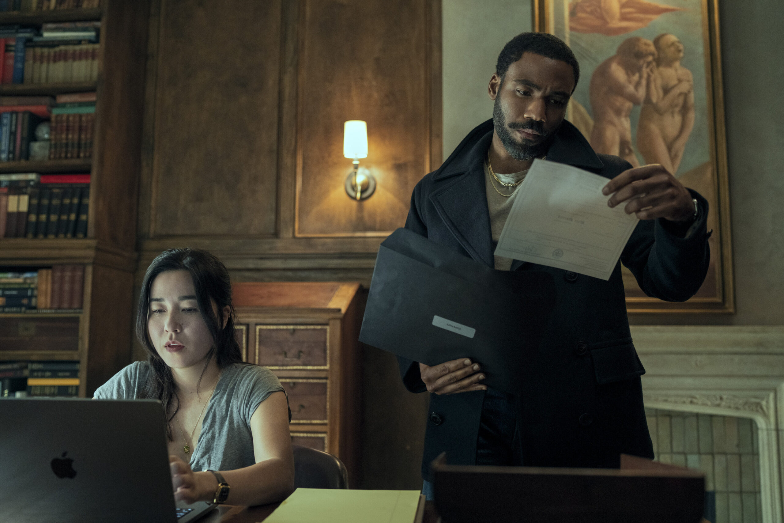 Maya Erskine as Mrs. Smith sitting at a desk in front of an Apple laptop and Donald Glover as Mr. Smith standing next to her holding a folder and looking at papers in the Prime Video series Mr. and Mrs. Smith.