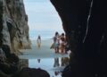 A long shot of a beach from inside a cave with a little girl staring forward and a group of young girls off to the side from the movie Seagrass.