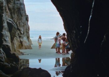 A long shot of a beach from inside a cave with a little girl staring forward and a group of young girls off to the side from the movie Seagrass.