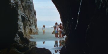A long shot of a beach from inside a cave with a little girl staring forward and a group of young girls off to the side from the movie Seagrass.