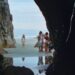 A long shot of a beach from inside a cave with a little girl staring forward and a group of young girls off to the side from the movie Seagrass.