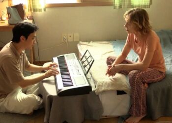 Ha Seong-guk as Inguk sitting on the floor playing a keyboard across from Isabelle Huppert as Iris sitting on a bed in the movie A Traveler's Needs.