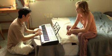 Ha Seong-guk as Inguk sitting on the floor playing a keyboard across from Isabelle Huppert as Iris sitting on a bed in the movie A Traveler's Needs.