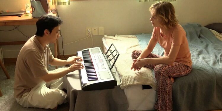 Ha Seong-guk as Inguk sitting on the floor playing a keyboard across from Isabelle Huppert as Iris sitting on a bed in the movie A Traveler's Needs.