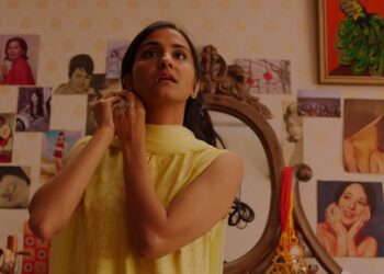 Amrit Kaur as Azra putting on an earring wearing a yellow dress in the movie The Queen of My Dreams.