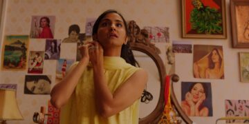 Amrit Kaur as Azra putting on an earring wearing a yellow dress in the movie The Queen of My Dreams.