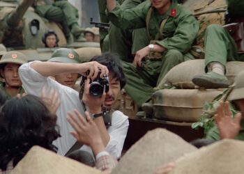 George Lam as Shiomi Akutagawa taking a photo around soldiers sitting on tanks parading through the streets in Boat People.