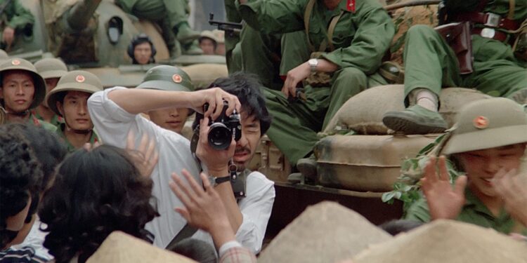 George Lam as Shiomi Akutagawa taking a photo around soldiers sitting on tanks parading through the streets in Boat People.