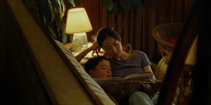 Choi Seung-yoon as So-young reading a book to Ethan Hwang as Dong-hyun on a couch in the movie Riceboy Sleeps.