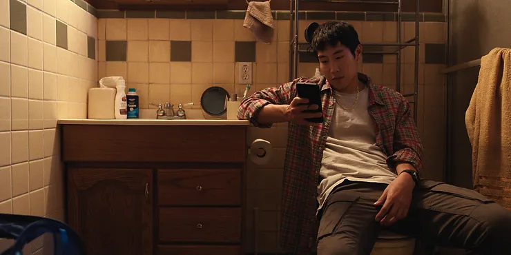 Young Mazino as Paul sitting on the toilet seat on his phone in the Netflix series BEEF.