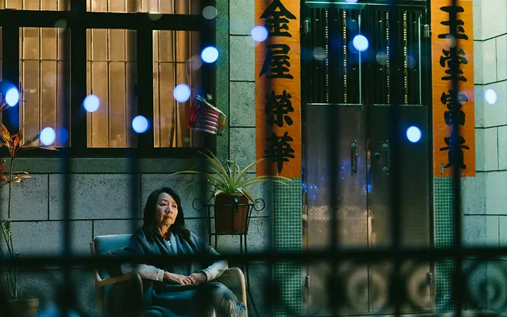Patra Au as Angie sitting on a chair outside a building in the Hong Kong movie All Shall Be Well.