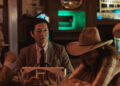 Jun Kunimura as Wada and Arata Iura as Hideki seated at a table in a cowboy bar opposite Robin Weigert as Peg in Tokyo Cowboy.