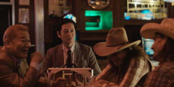 Jun Kunimura as Wada and Arata Iura as Hideki seated at a table in a cowboy bar opposite Robin Weigert as Peg in Tokyo Cowboy.
