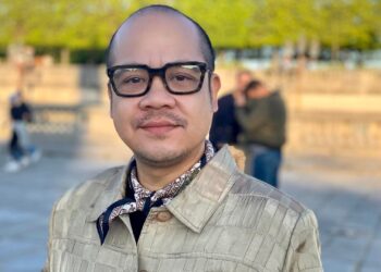 A medium close-up portrait of Jeremiah Abraham, a Filipino man wearing black glasses and a tan jacket.