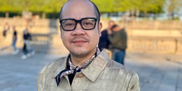 A medium close-up portrait of Jeremiah Abraham, a Filipino man wearing black glasses and a tan jacket.