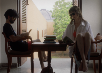 Sathya Sridharan as Ben and Anastasia Olowin as Suzanne sitting on wooden chair across from each other with a matching table with books piled on top in between them in "Ben and Suzanne, A Reunion In Four Parts".