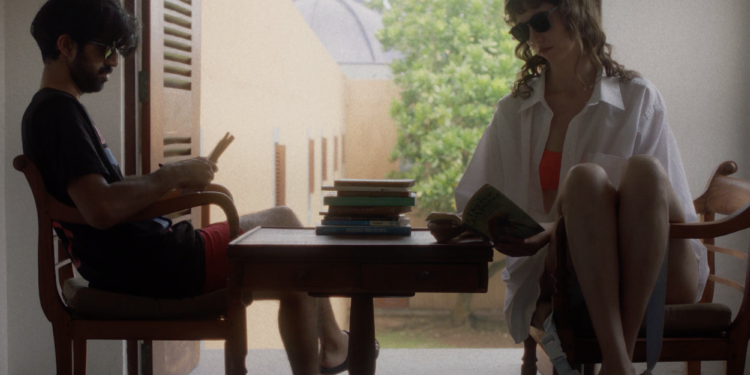 Sathya Sridharan as Ben and Anastasia Olowin as Suzanne sitting on wooden chair across from each other with a matching table with books piled on top in between them in "Ben and Suzanne, A Reunion In Four Parts".