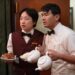Jimmy O. Yang as Willis Wu and Ronny Chieng as Fatty Choi holding plates and tea pots in the television series Interior Chinatown.
