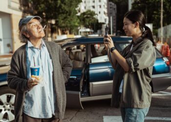 Standing in front of a blue car on the street, Jennifer Yu as Kay takes the picture of David Chiang in 'In Broad Daylight.'