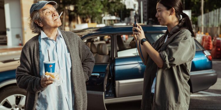 Standing in front of a blue car on the street, Jennifer Yu as Kay takes the picture of David Chiang in 'In Broad Daylight.'