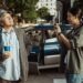 Standing in front of a blue car on the street, Jennifer Yu as Kay takes the picture of David Chiang in 'In Broad Daylight.'