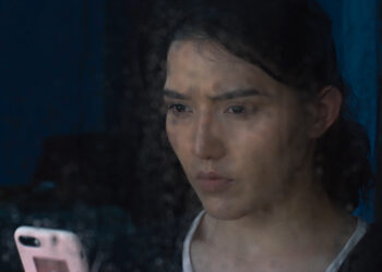 Guzalnur Uchqun in Nikah staring out a window on a rainy day holding a mobile phone.