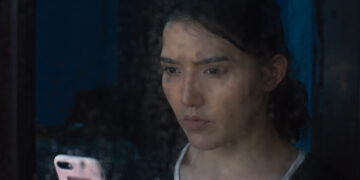 Guzalnur Uchqun in Nikah staring out a window on a rainy day holding a mobile phone.