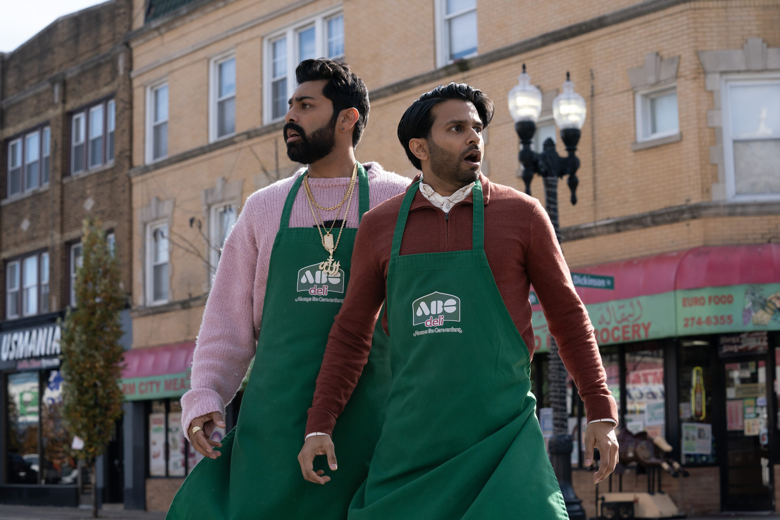 Saagar Shaikh as Raj Dar and Asif Ali as Mir Dar standing on the street wearing green aprons that say ABC Deli looking shocked in the TV series Deli Boys.