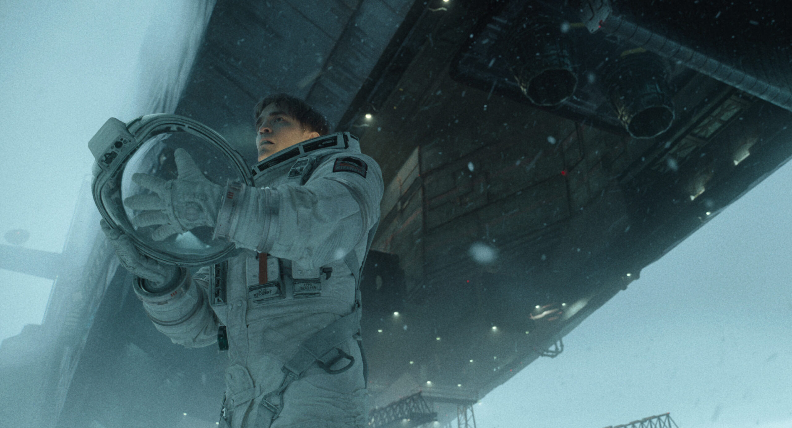 Robert Pattinson as Mickey Barnes in a space suit, holding his helmet in a snowy landscape in Mickey 17.