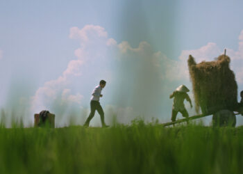 A still form the documentary Ba's Book of a man in a field walking towards two workers lifting a bale of hay